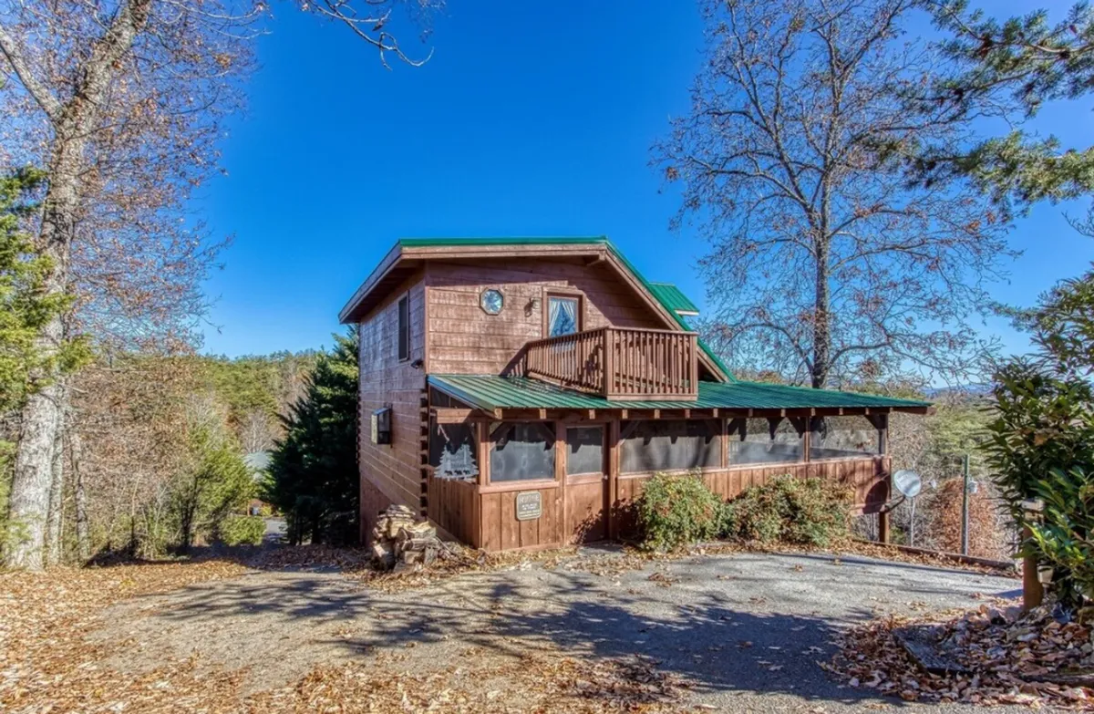 This image captures the wooden exterior of the house with a green metal roof, a spacious balcony with wooden railing, surrounding trees, and a driveway leading up to the cabin. The inviting setting hints at outdoor activities and gathering spots, surrounded by the natural beauty of the Smoky Mountains.