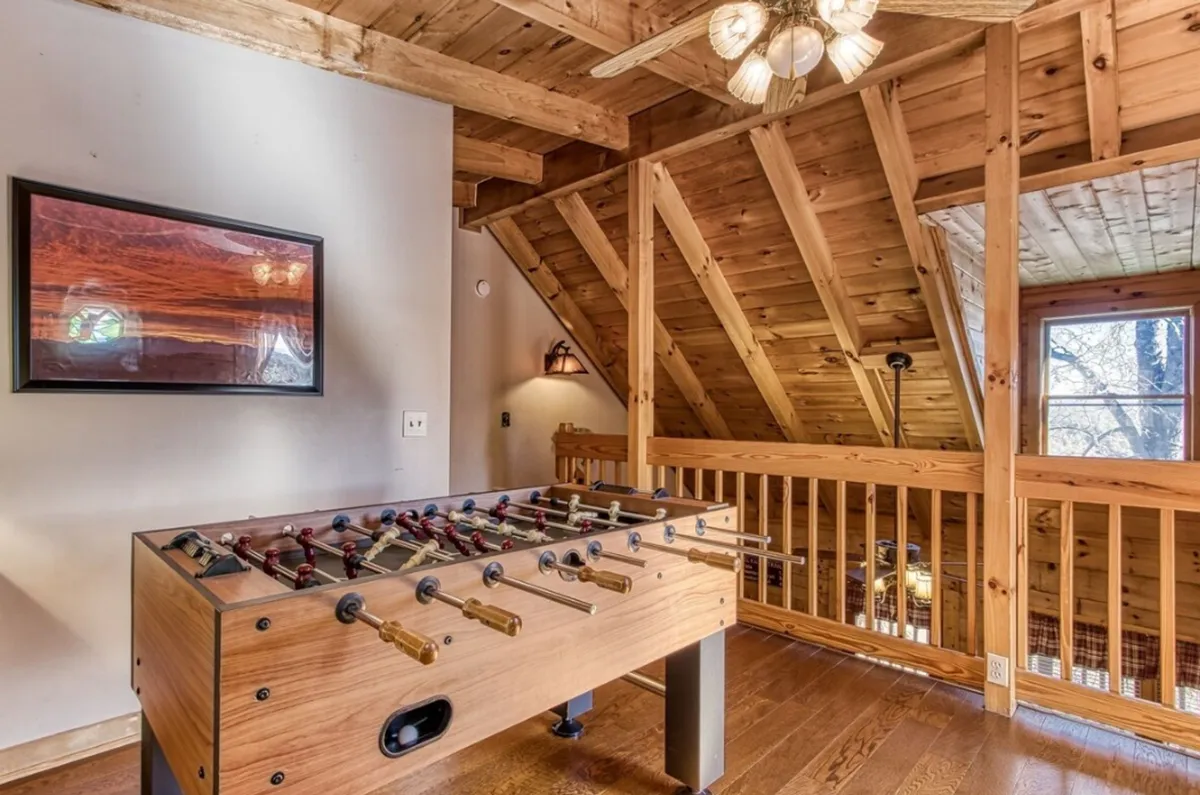 A cozy, well-lit game room featuring a foosball table under rustic wooden beams, with a large window letting in natural light and framed artwork adding a homey touch. The space is arranged for socializing and fun.
