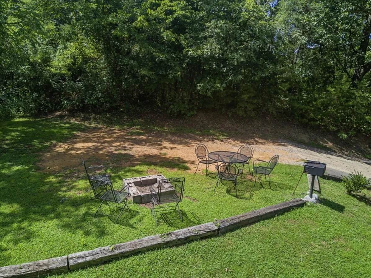 An inviting exterior setting with a fire pit surrounded by comfortable outdoor seating, a charcoal grill nearby, and lush greenery creating a natural backdrop. The scene suggests the warmth of gathering around the fire and opportunities for barbecues and al fresco dining.