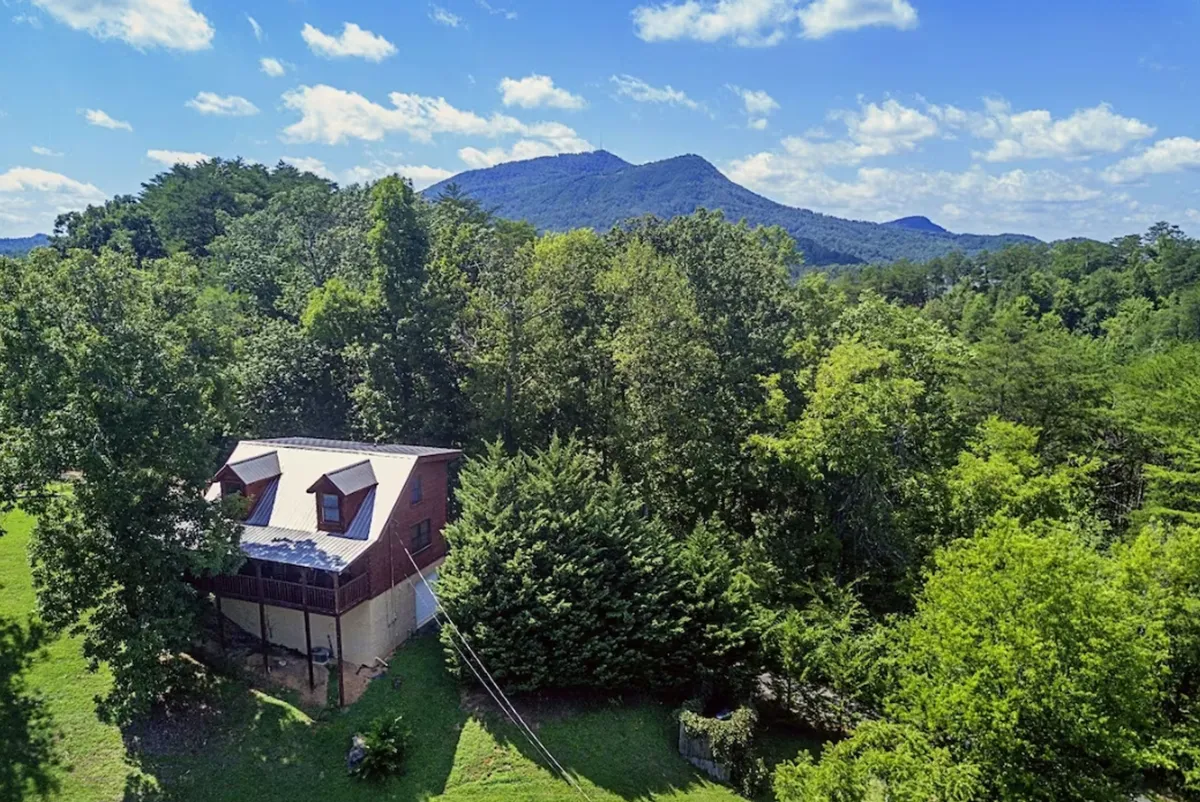 A wide view of the house nestled among trees with a stunning mountain backdrop. The scene invokes the tranquility of mountain mornings and outdoor fun, with hints of hiking, wildlife sightings, and cozy evenings by the fire pit.