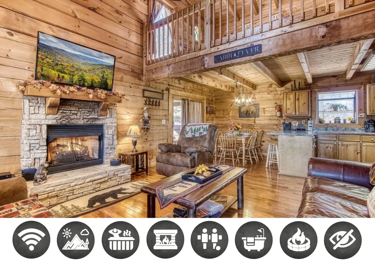 The image captures the cozy heart of the cabin's open-concept living space, featuring a crackling stone fireplace, inviting sofa, rustic dining table set for gatherings, and a well-equipped kitchen—all bathed in the natural light of the Smoky Mountains.