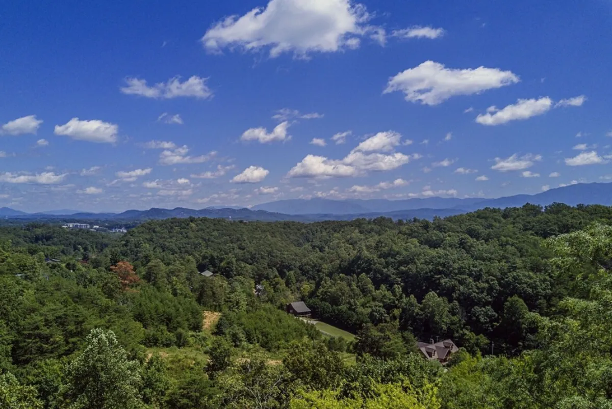 A wide, open landscape stretches out with lush green fields, distant mountains, and a brilliant blue sky dotted with clouds. The view promises adventure and a natural playground for hiking, photography, or simply savoring the scenery with friends.