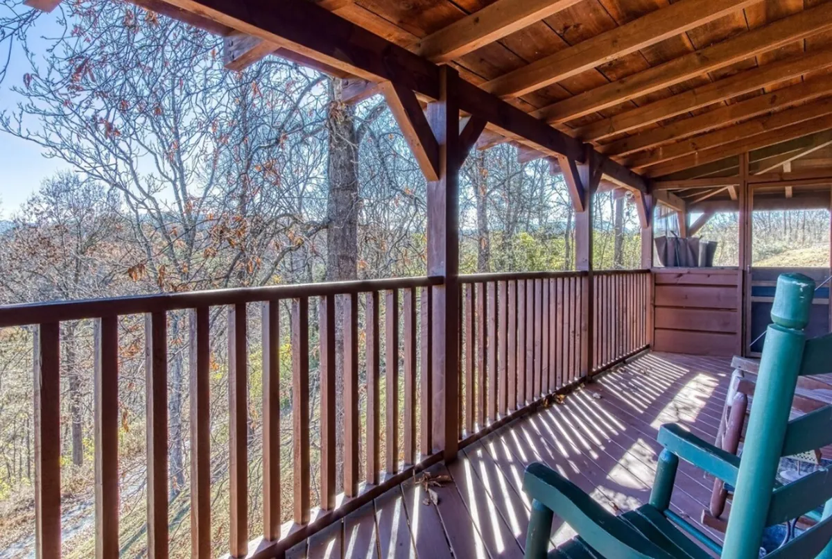 An inviting exterior shot featuring a wooden deck with a green rocking chair, surrounded by trees and framed by rustic wooden railings. The space promises a peaceful transition between the comforts of the cabin and the beauty of the outdoors, perfect for relaxing after a day of exploration.