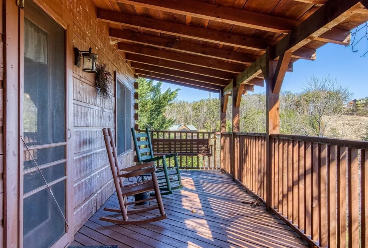 The image features rocking chairs and a swing bench on a wooden porch surrounded by lush natural scenery. The atmosphere is serene and inviting, perfect for relaxing with a partner, sharing quiet conversation, or enjoying the sounds of nature.
