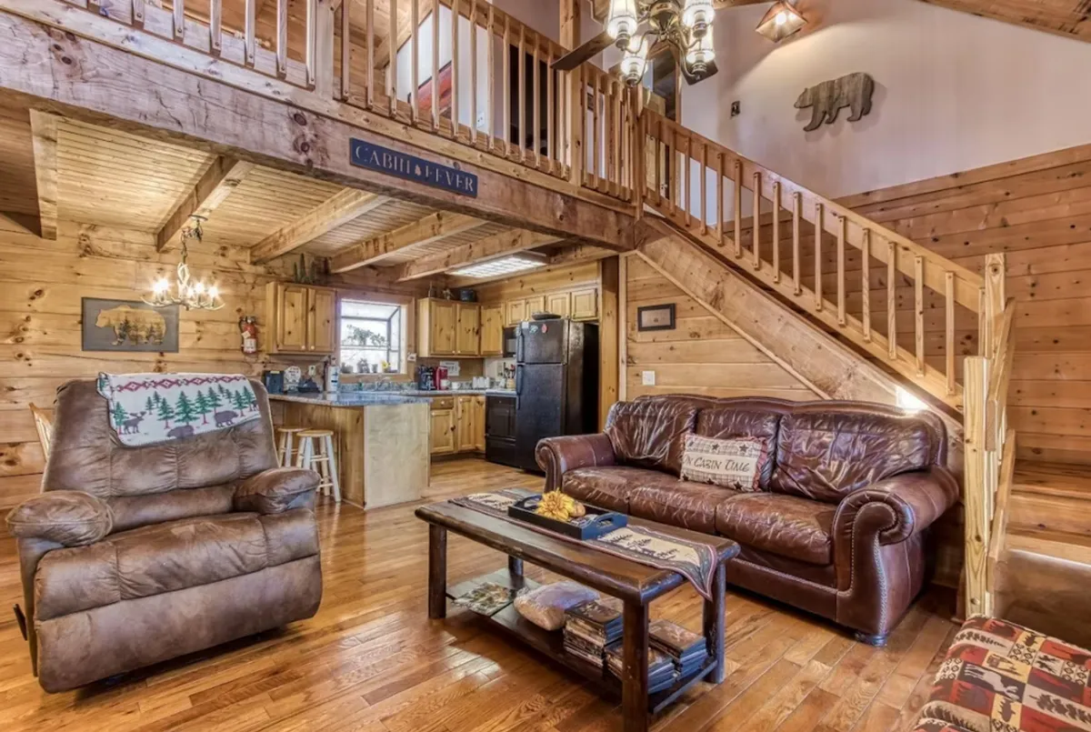 The image shows a warm, open-plan living area with a leather sofa, wooden staircase, chandelier, and a clear view of the kitchen. The space is bathed in soft light and invites guests to gather, relax, and seamlessly move between lounging and meal preparation.