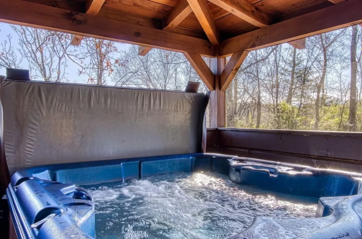 A warmly lit interior scene featuring a hot tub with exposed wooden beams and abundant natural light streaming in. The view from the hot tub suggests a relaxing environment perfect for unwinding after a day out.