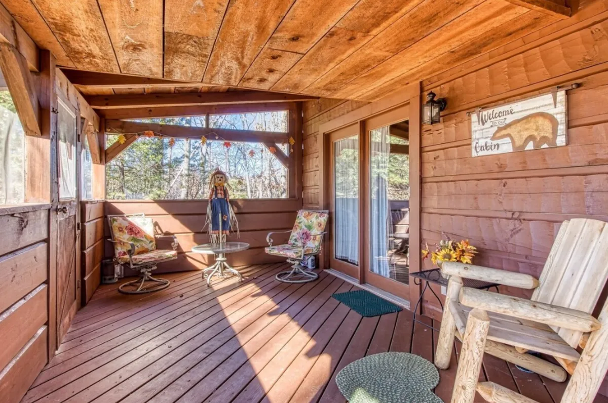 A cozy, partially sheltered seating area with wooden chairs, a table adorned with flowers, and a welcoming seasonal scarecrow. Trees are visible just beyond, blending indoor comfort with outdoor ambiance—ideal for sharing meals or enjoying the sights and sounds of nature.