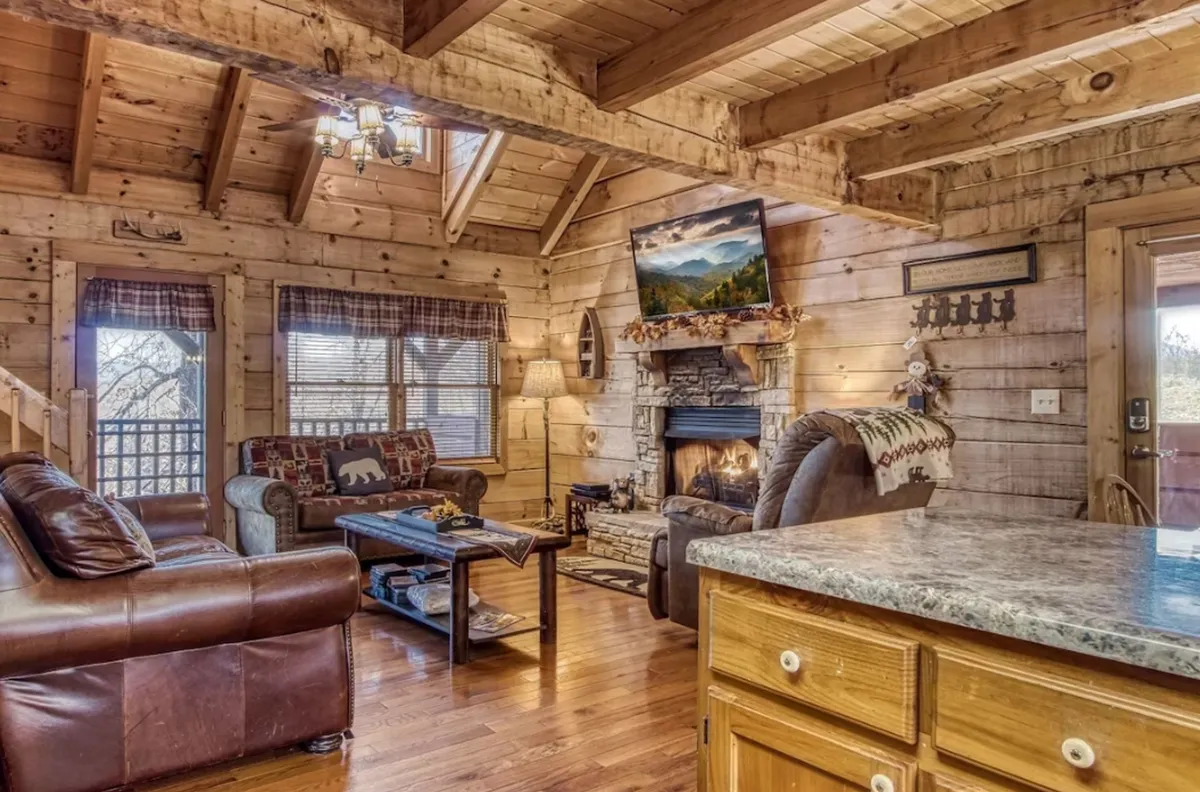 This image captures a wide view of the cabin's main living space, highlighting the cozy fireplace, a plush sofa, rustic wooden beams overhead, a television, and a farmhouse dining table all within one inviting room. Warm lighting and natural wood elements convey a welcoming and communal atmosphere.