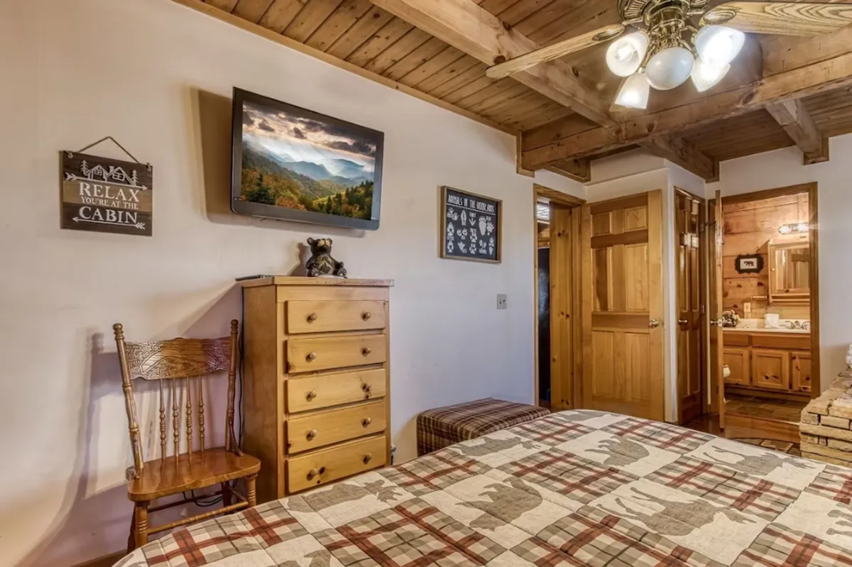 The image highlights a king bedroom with a plaid bedspread, wooden furniture, a TV displaying a landscape, and a decorative bear figurine. The warm lighting and nature views from the window evoke comfort and relaxation.
