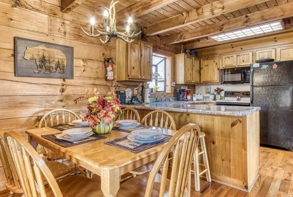 A warmly lit dining area with a large wooden farmhouse table sits at the center, flanked by a chandelier, bear-themed artwork, and the kitchen's countertop in view. The space exudes rustic charm and is clearly designed for communal meals and celebrations.