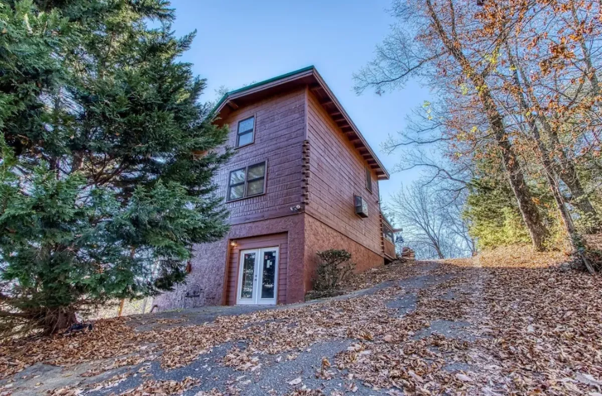The image shows the wooden exterior of the cabin nestled among tall, leafy trees with a spacious driveway leading up to the home. The porch invites guests to relax and soak in the surrounding nature, visually hinting at group gatherings and easy access to the outdoors.