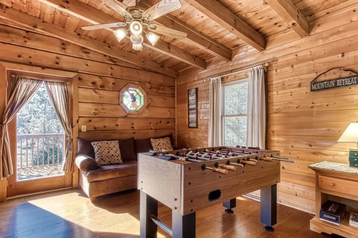 A bright, airy loft space featuring a foosball table, a cozy couch for lounging, a striking stained glass window, and exposed wooden beams. Natural light streams in, creating an inviting, playful setting perfect for gathering with friends and family.