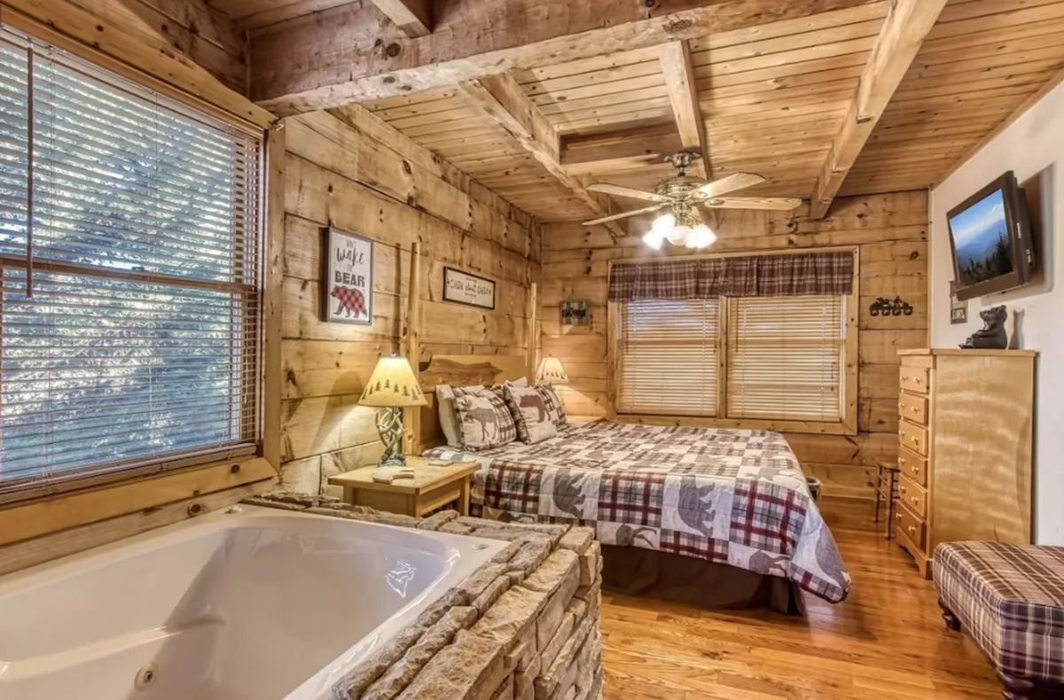 This image features a warm, rustic bedroom with wooden walls, a king-sized bed adorned in plaid bedding, glowing lamps, and a large jacuzzi tub right in the suite. Natural light streams in, creating a cozy, inviting atmosphere.